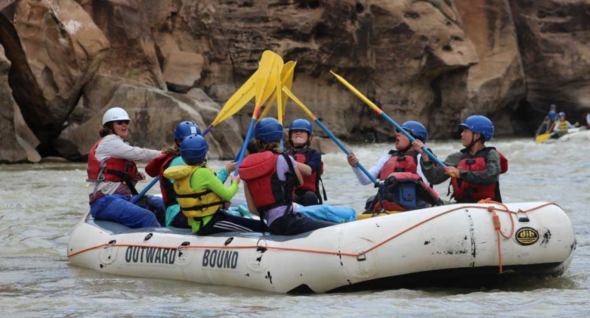 On board a raft, people wearing helmets and life jackets raise their oars together. 